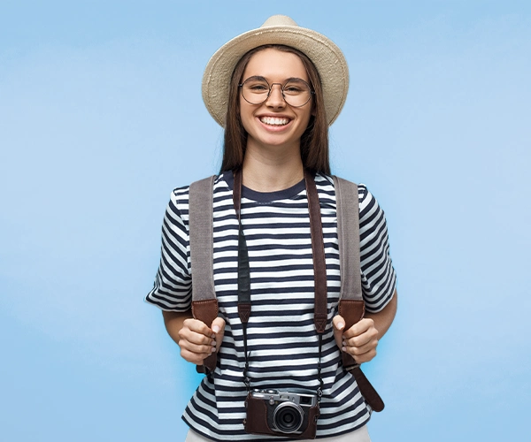 Gut vorbereitet auf Reisen Junge Frau mit Rucksack, Sonnenhut und Kamera vor blauem Hintergrund – symbolisiert eine gesunde und gut vorbereitete Reisende.
