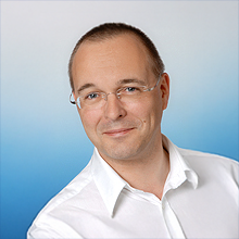 Dr. med Thomas Schwarz Porträt eines lächelnden Mannes mit Brille in weißem Hemd vor blau-weißem Hintergrund – dargestellt ist der Praxisinhaber Dr. med. Thomas Schwarz.