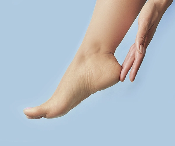 Nahaufnahme einer Hand, die eine Ferse eines ausgestreckten nackten Fußes berührt, vor hellblauem Hintergrund.