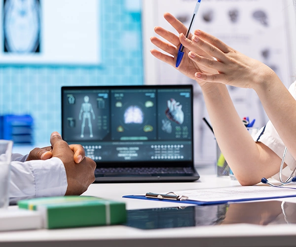 Ärztliches Beratungsgespräch in der Inneren Medizin mit zwei Personen am Schreibtisch, medizinischen Befunden auf einem Laptop und erklärenden Handgesten zur Diagnose von internistischen Erkrankungen.